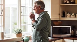 Ein älterer Mann steht in der Küche am Fenster und trinkt einen Tee gegen seine Prostata-Beschwerden.