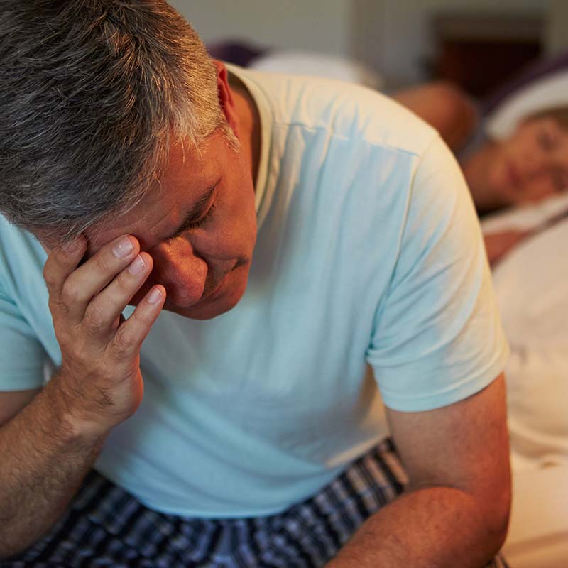 Mann sitzt wegen nächtlichem Harndrang wach auf der Bettkante, während seine Frau im Hintergrund schläft.